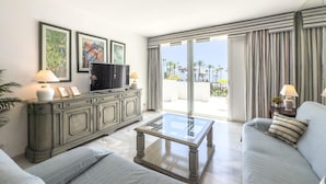 A modern living room with a large flat-screen TV, a glass coffee table, and a sofa. There are framed pictures on the wall and a view of palm trees outside.