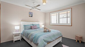 A bedroom with a bed, a nightstand, a lamp, a ceiling fan, a window with blinds, and a small stool. - Akuli By The Sea Unit 2 (Iluka)