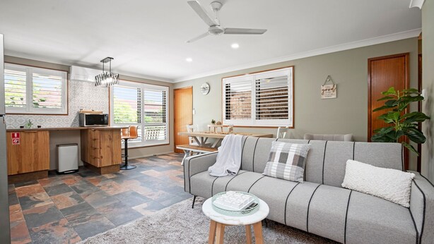 A modern living room with a grey sofa, a small round table, and a kitchen area with a microwave and a sink. - Akuli By The Sea Unit 2 (Iluka)