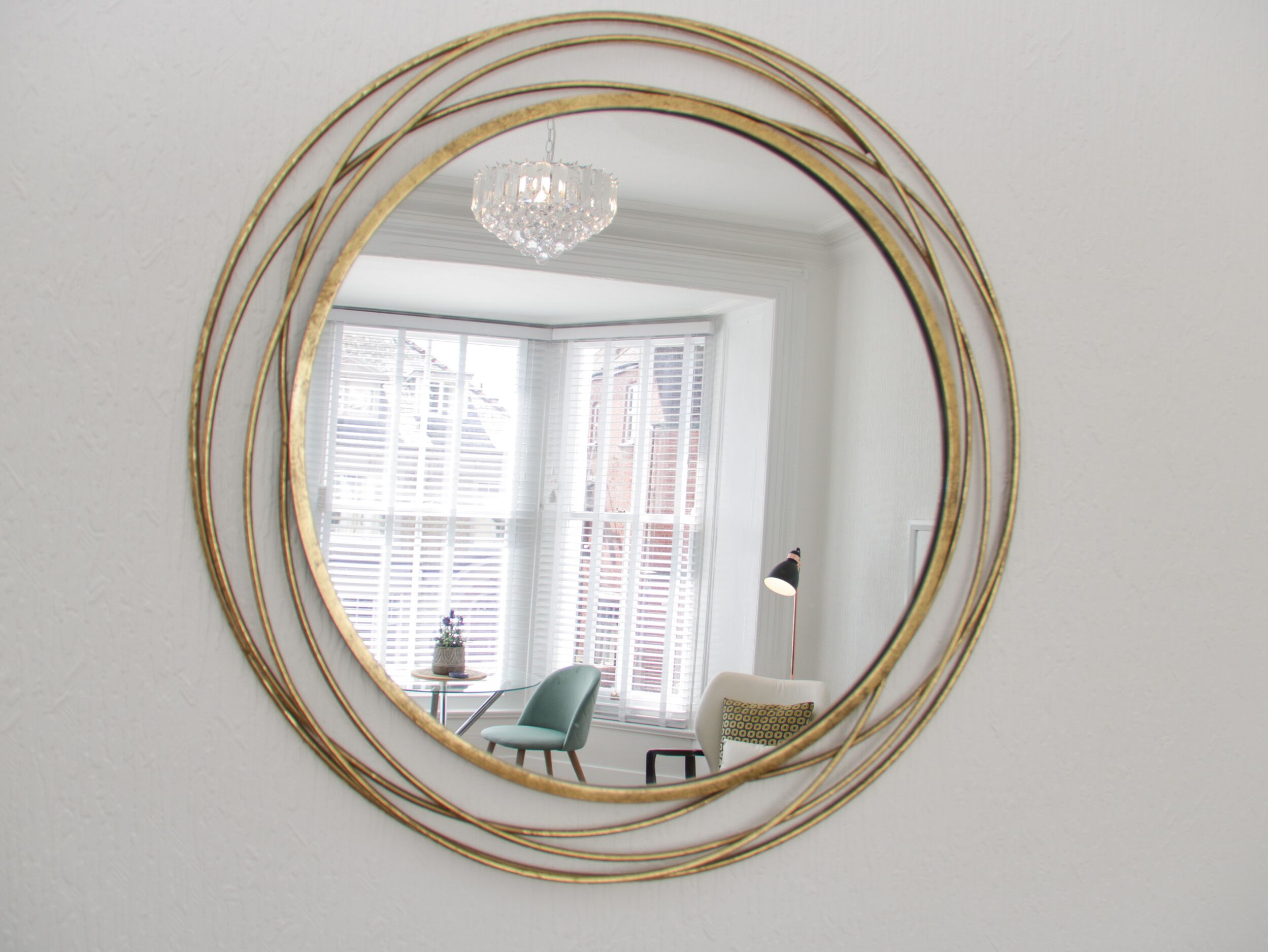 A room with a large oval mirror on the wall, a chandelier, a window with blinds, and a chair.