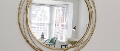 A room with a large oval mirror on the wall, a chandelier, a window with blinds, and a chair.