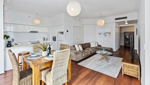 Une cuisine moderne avec des meubles blancs, un coin repas avec des chaises en osier, un canapé gris et un téléviseur dans le salon.