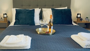 Un lit bien fait, recouvert d’une couverture bleu foncé, avec deux oreillers, un plateau sur lequel se trouvent une bouteille et des verres, et un tableau représentant des voiliers accroché au mur.