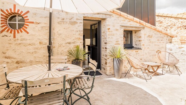 Un patio con una mesa redonda de madera y sillas, un parasol grande y plantas en macetas.