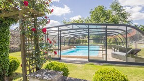 Una piscina cubierta con paredes de vidrio y techo curvo, rodeada de vegetación y un muro de piedra.