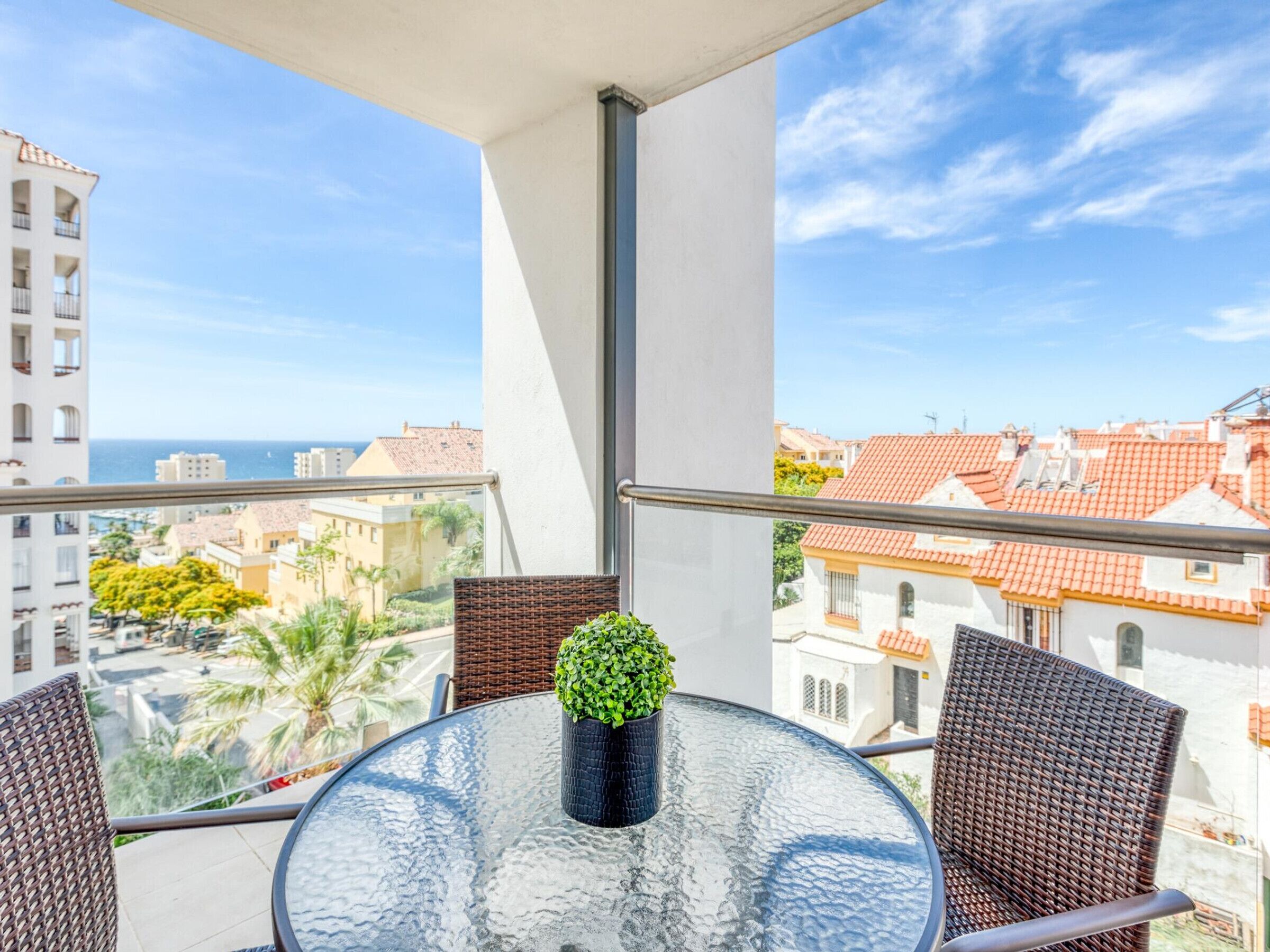 A balcony with a glass table, wicker chairs, and a potted plant.