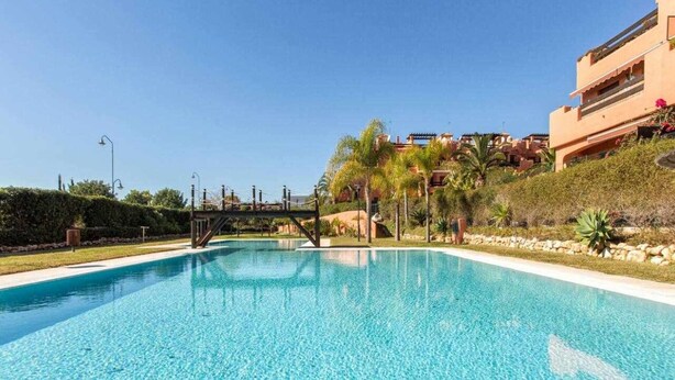Una piscina azul cristalina con un puente, rodeada de vegetación y edificios.