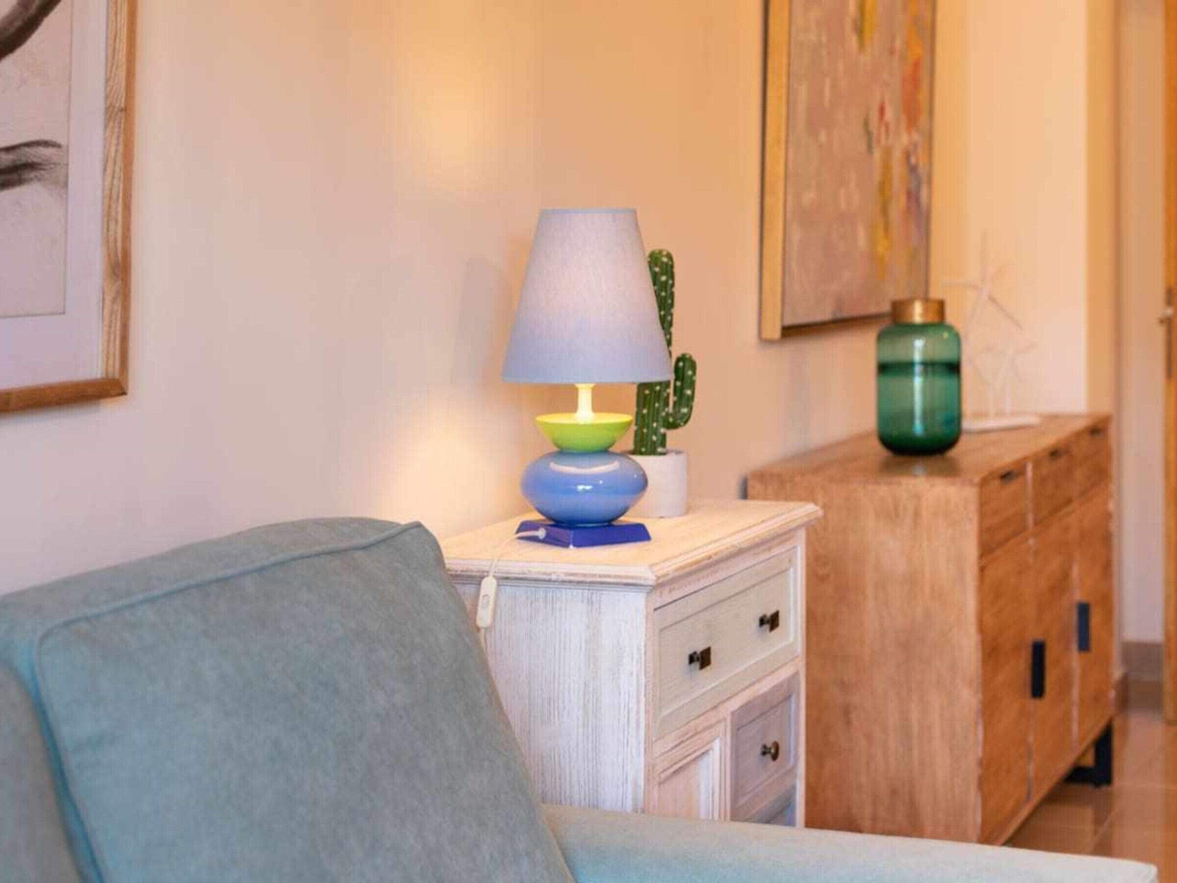 Ein Wohnzimmer mit einem blauen Sofa, einer bunten Lampe, einem Kaktus und einem Holzschrank.