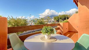 Mesa redonda con una planta en maceta en el centro, rodeada por dos sillas verdes, ubicada en un balcón con vistas a un paisaje urbano y montañas al fondo.