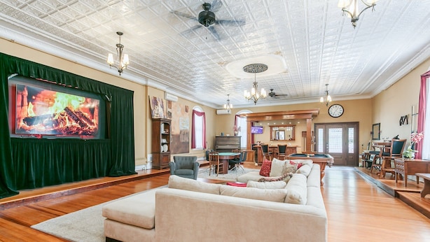 A spacious living room with a large flat-screen TV, a beige sofa, wooden flooring, and a ceiling with intricate patterns.