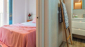 Quarto com cama, mesa de cabeceira com um vaso de flores e banheiro com espelho e porta-toalhas.