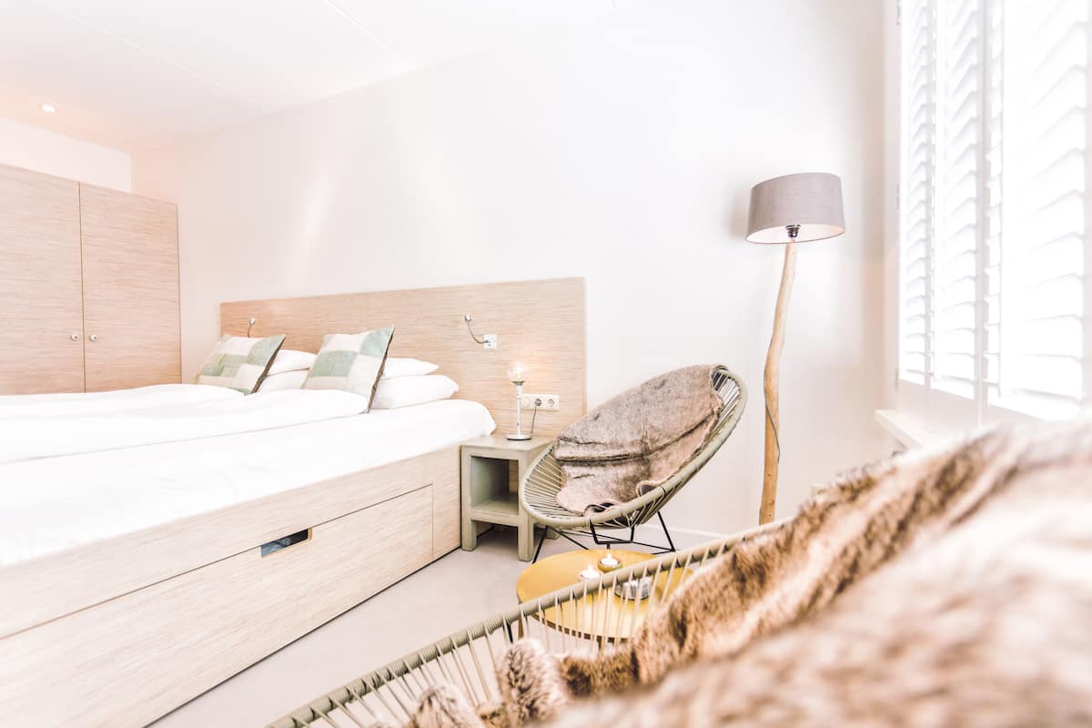 A modern bedroom with a large bed, bedside table, chair, lamp, and window with blinds.