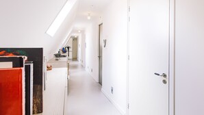 A bright, narrow hallway with white walls and a sloped ceiling. There are doors on the right, and a radiator on the left. - Logement Overzee (Nes)