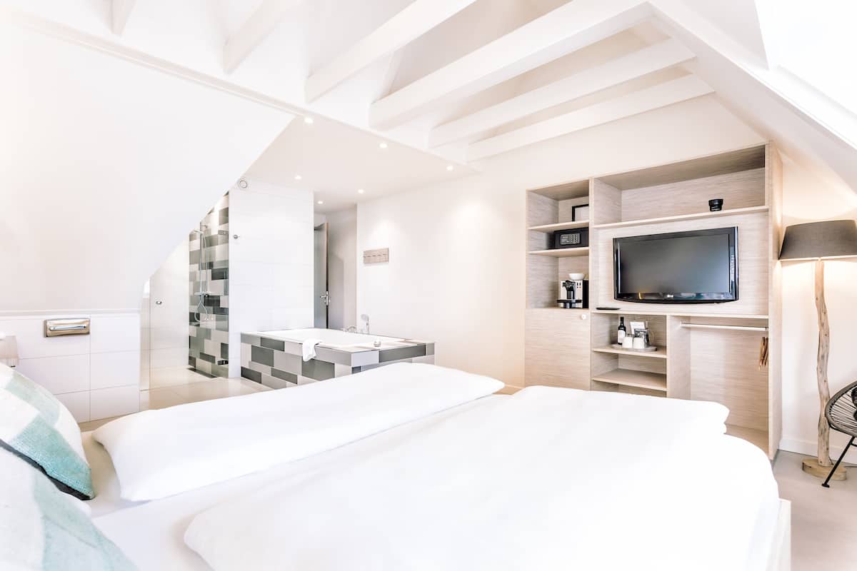 A modern hotel room with a large bed, a TV, a built-in shower, and a small seating area.
