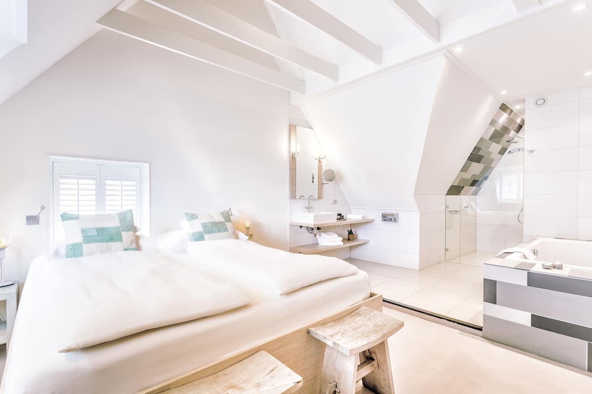 A modern bedroom with a large bed, a bedside table, a wooden bench, a bathroom with a bathtub, a shower, and a toilet.