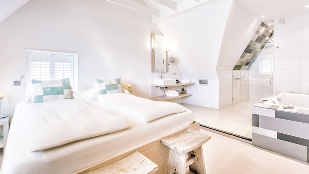 A modern bedroom with a large bed, a wooden bench, a bathroom with a bathtub, and a staircase. - Logement Overzee (Nes)