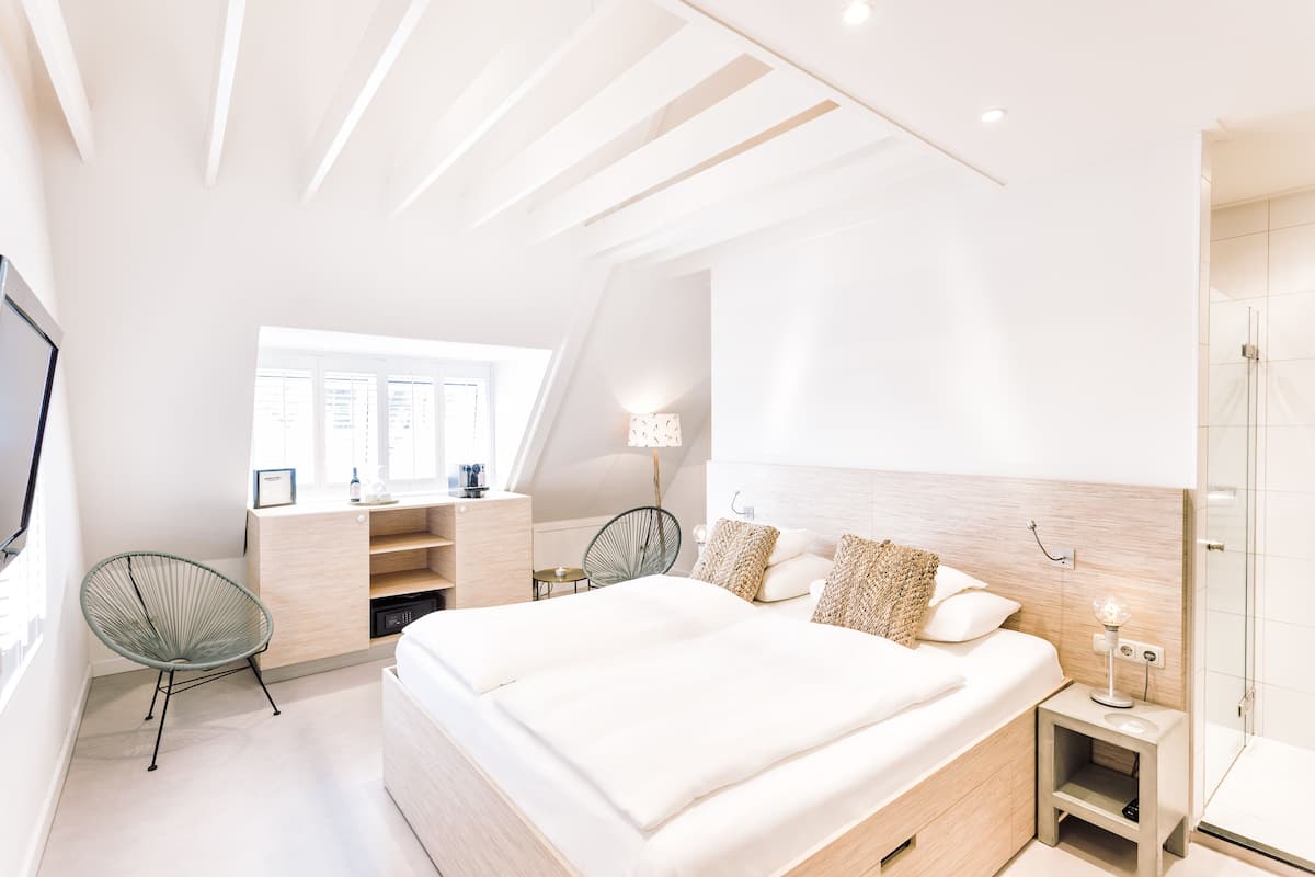 A modern bedroom with a large bed, a desk with a chair, a small table with a lamp, and a shower area.