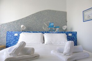 Habitación de hotel con cama, ropa blanca, dos toallas blancas, cabecera de mosaico azul y blanco, y una silla pequeña azul y blanca.