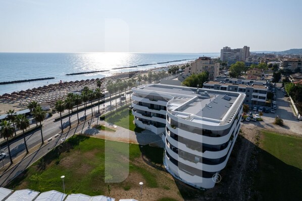 Un immeuble moderne à plusieurs étages, doté d’un toit plat, situé près d’une plage, d’une promenade et de palmiers.