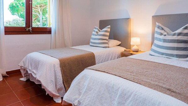 Une chambre d’hôtel avec deux lits, chacun agrémenté d’un oreiller à rayures et d’une tête de lit. Il y a une fenêtre donnant sur des arbres, ainsi qu’une table de chevet avec une lampe placée entre les deux lits.