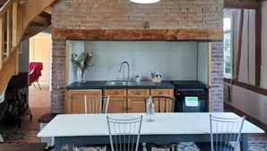 A kitchen with a brick wall, wooden beams, a hanging light fixture, a sink, cabinets, and a dining table with chairs.
