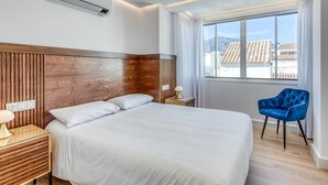 Une chambre d’hôtel moderne avec un grand lit, une tête de lit en bois, une fenêtre donnant sur des immeubles et une chaise bleue.