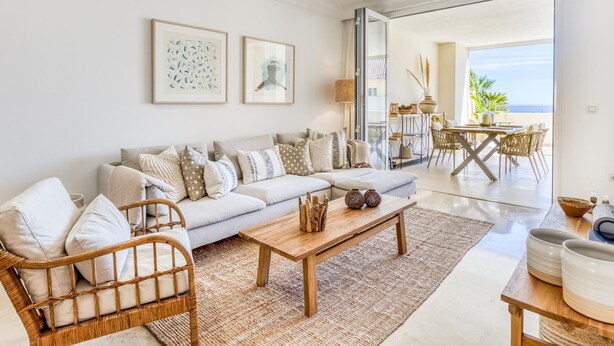A spacious living room with a sofa, a wooden coffee table, and a wicker chair. There is a dining area with a table and chairs, and a view of the ocean through large windows. - Sun-Drenched Apartment in Estepona - 2BR, Pools, Near Beaches & Old Town (Estepona)