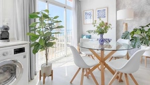 Ett modernt kök med tvättmaskin, en matplats med ett glasbord och vita stolar, och utsikt över omgivningarna genom stora fönster.