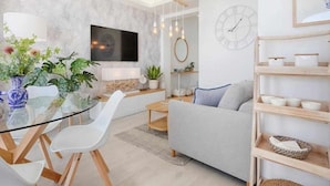 Ett modernt vardagsrum med ett glasbord, vita stolar, en grå soffa och en trähylla med dekorativa föremål.