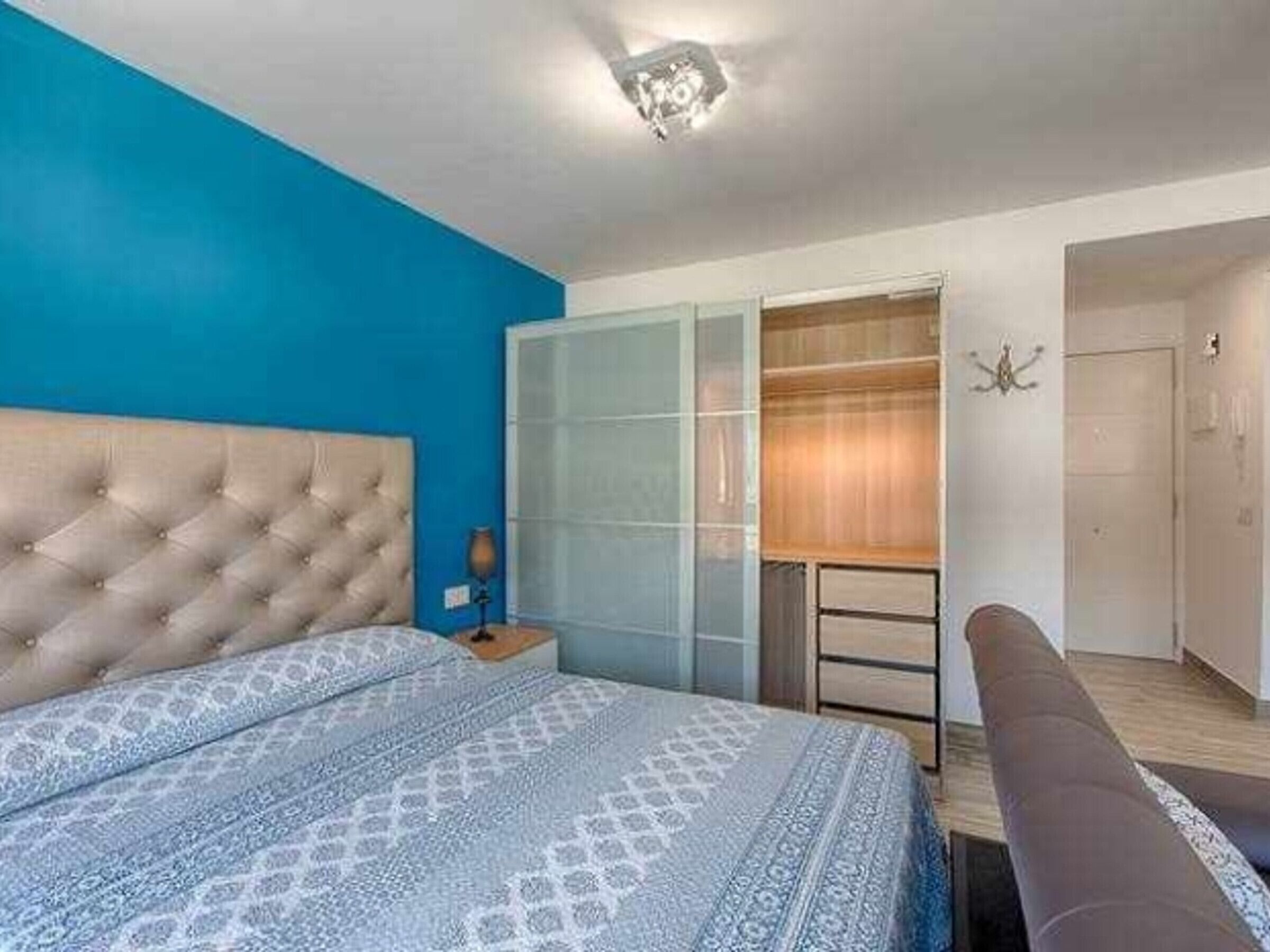 A bedroom with a tufted headboard, a patterned bedspread, a wardrobe with glass doors, and a bedside table with a lamp.