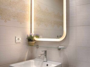 Une salle de bain avec un lavabo blanc, un miroir avec un cadre doré et une petite étagère sur laquelle est posée une plante.