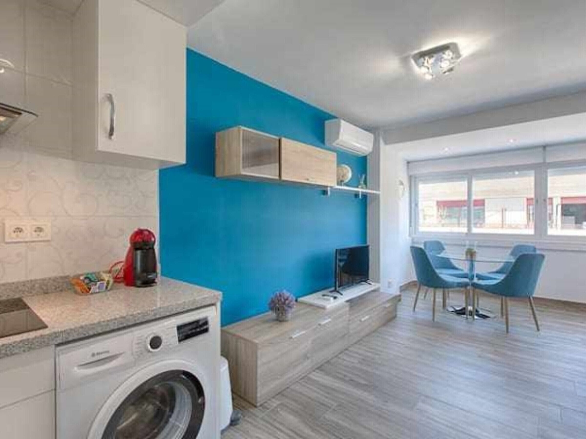 A modern kitchen with a washing machine, a blue accent wall, and a dining area with blue chairs.