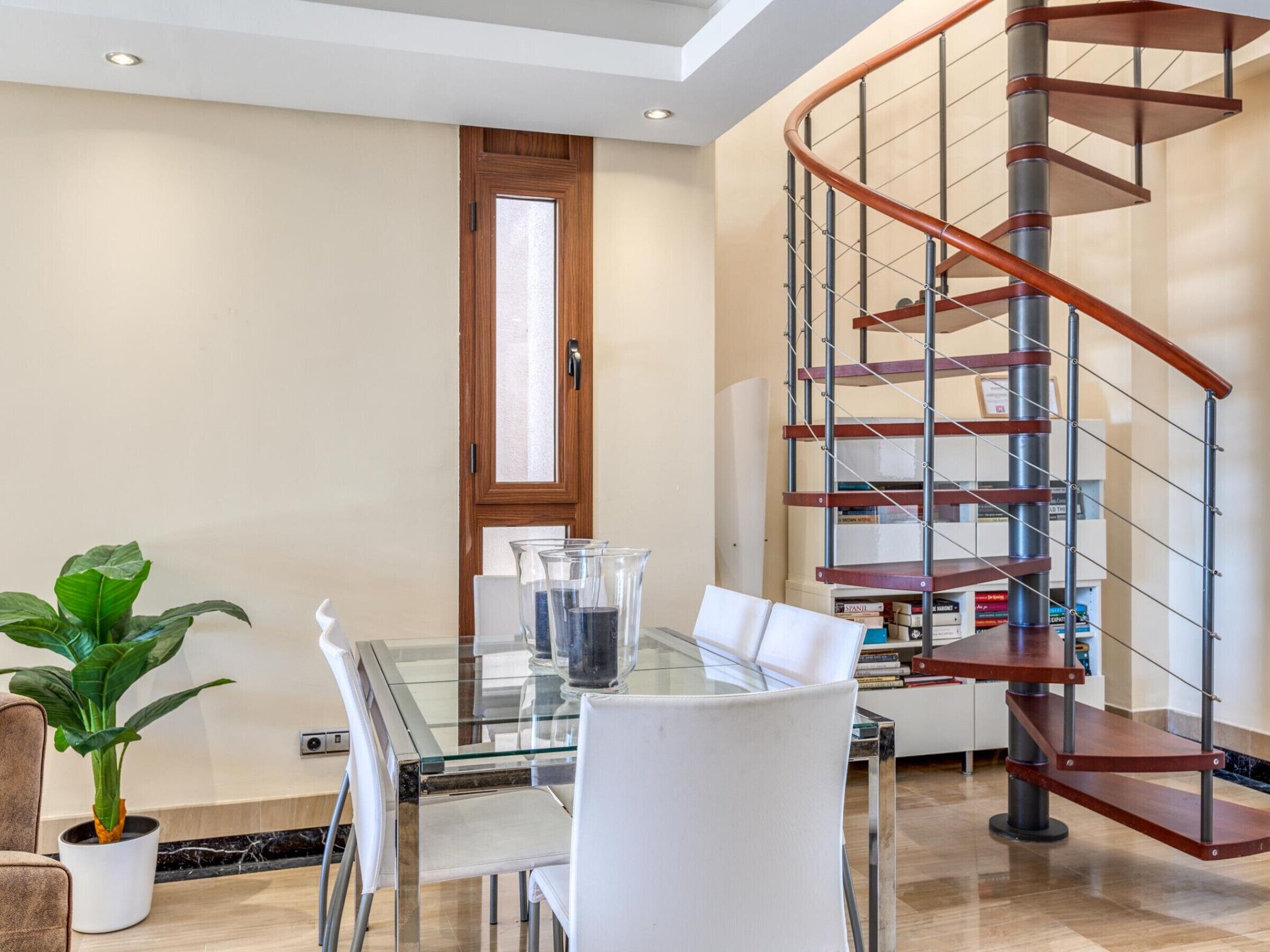 A modern interior with a glass dining table, white chairs, a potted plant, and a spiral staircase.