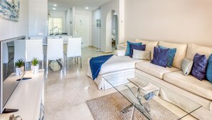 Una sala de estar moderna con una mesa de centro de vidrio, un sofá color beige con cojines azules y morados, un televisor de pantalla plana y un comedor con sillas blancas y una mesa de vidrio.