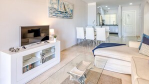 Una sala de estar moderna con televisor, muebles blancos, un comedor y una cocina al fondo.