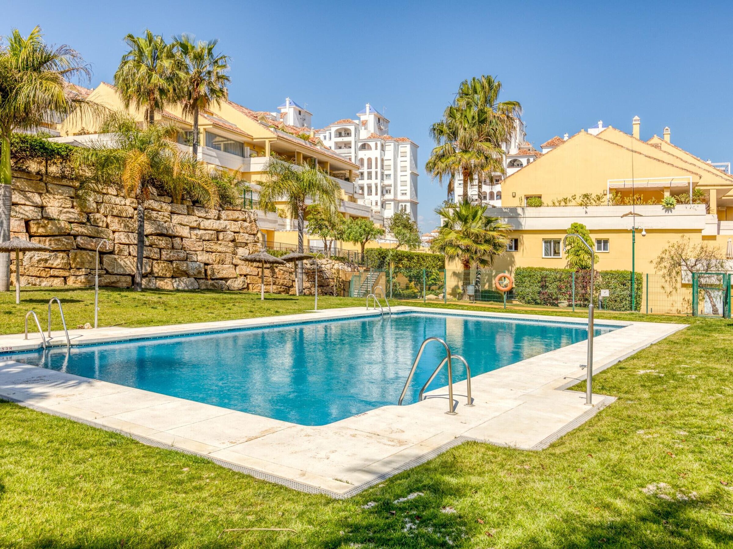 Una piscina con un muro de piedra y palmeras, rodeada de edificios con balcones.
