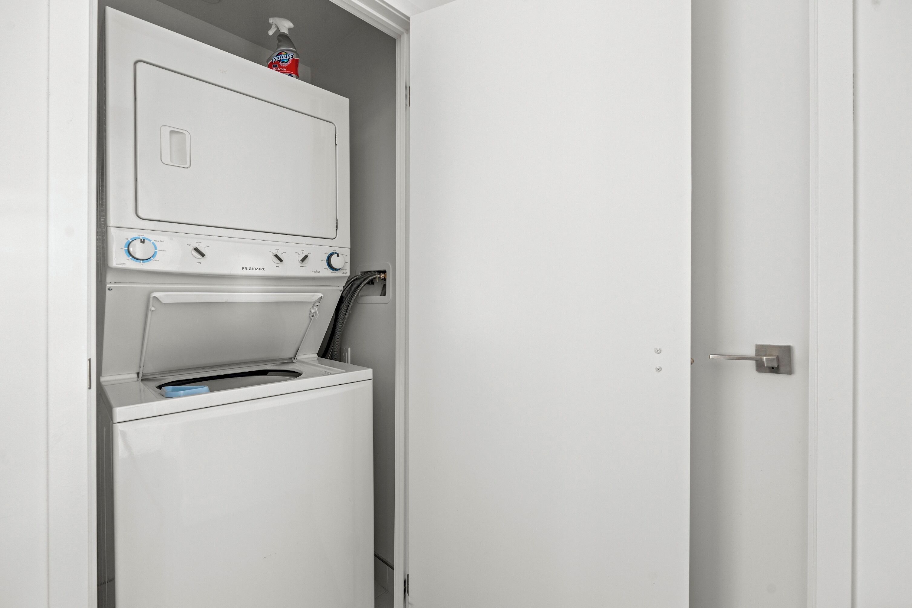 A compact, white laundry room with a front-loading washer and dryer, a control panel with various settings, and a small shelf with a bottle of detergent.