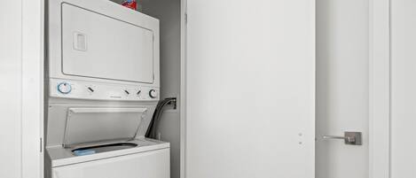 A compact, white laundry room with a front-loading washer and dryer, a control panel with various settings, and a small shelf with a bottle of detergent.