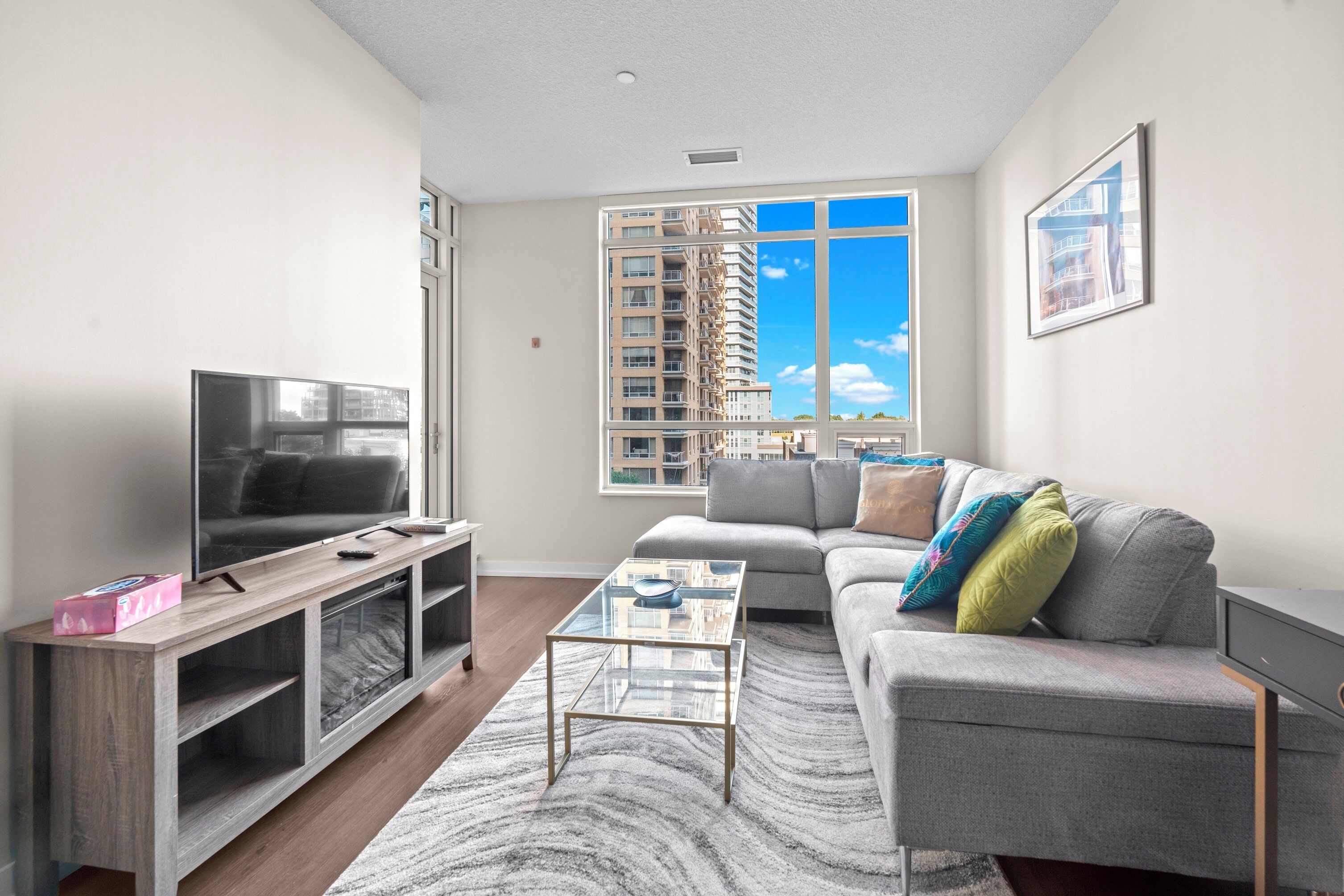 A modern living room with a flat-screen TV, a glass coffee table, a grey sofa with colorful cushions, and a large window offering a city view.