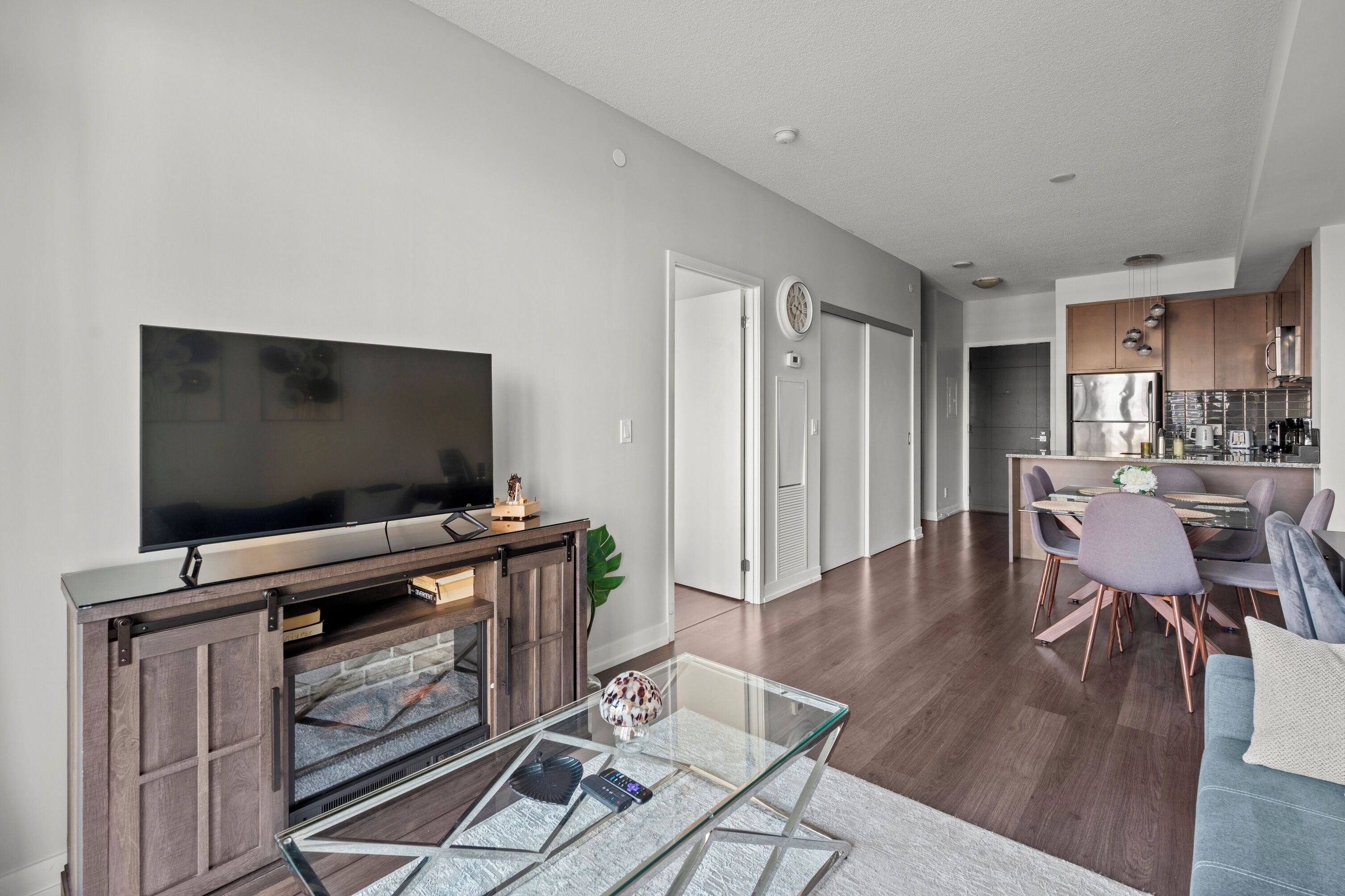A modern living room with a flat-screen TV, a glass coffee table, and a dining area with a table and chairs.