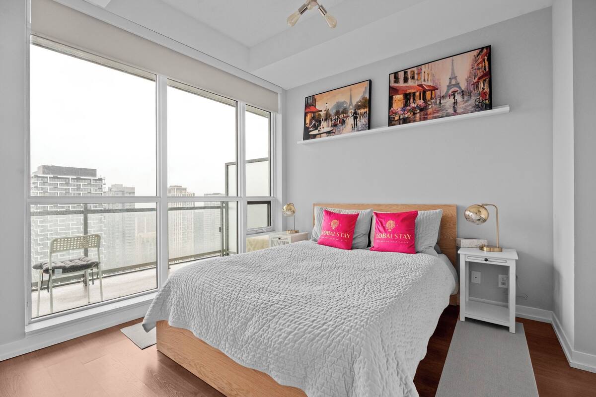 Ein Schlafzimmer mit einem großen Fenster, einem Bett mit rosa und grauen Kissen, einem Nachttisch mit einer Lampe und einem Stadtbild.