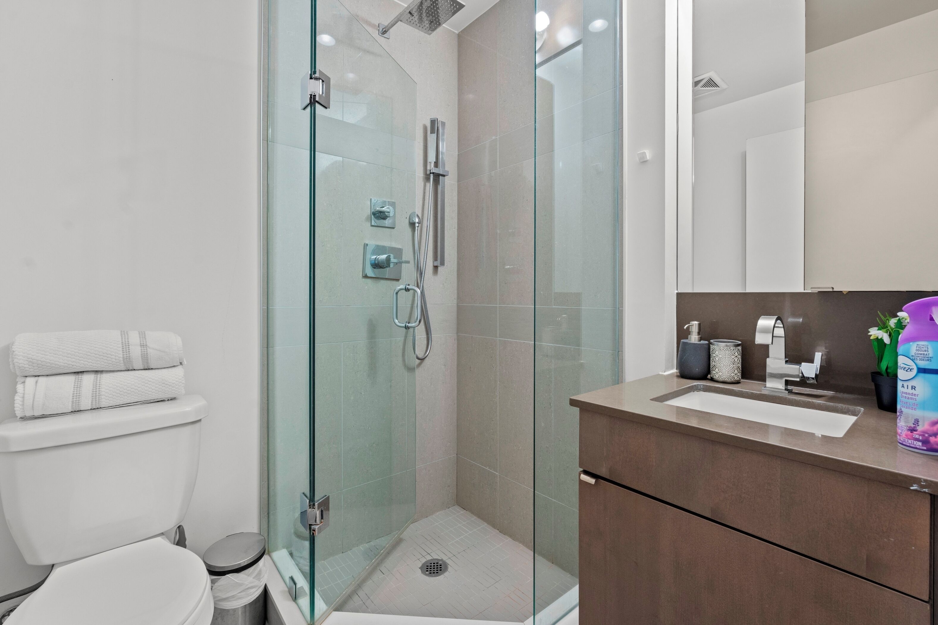 Un baño moderno con cabina de ducha de vidrio, inodoro, lavamanos con espejo y un cubo de basura.