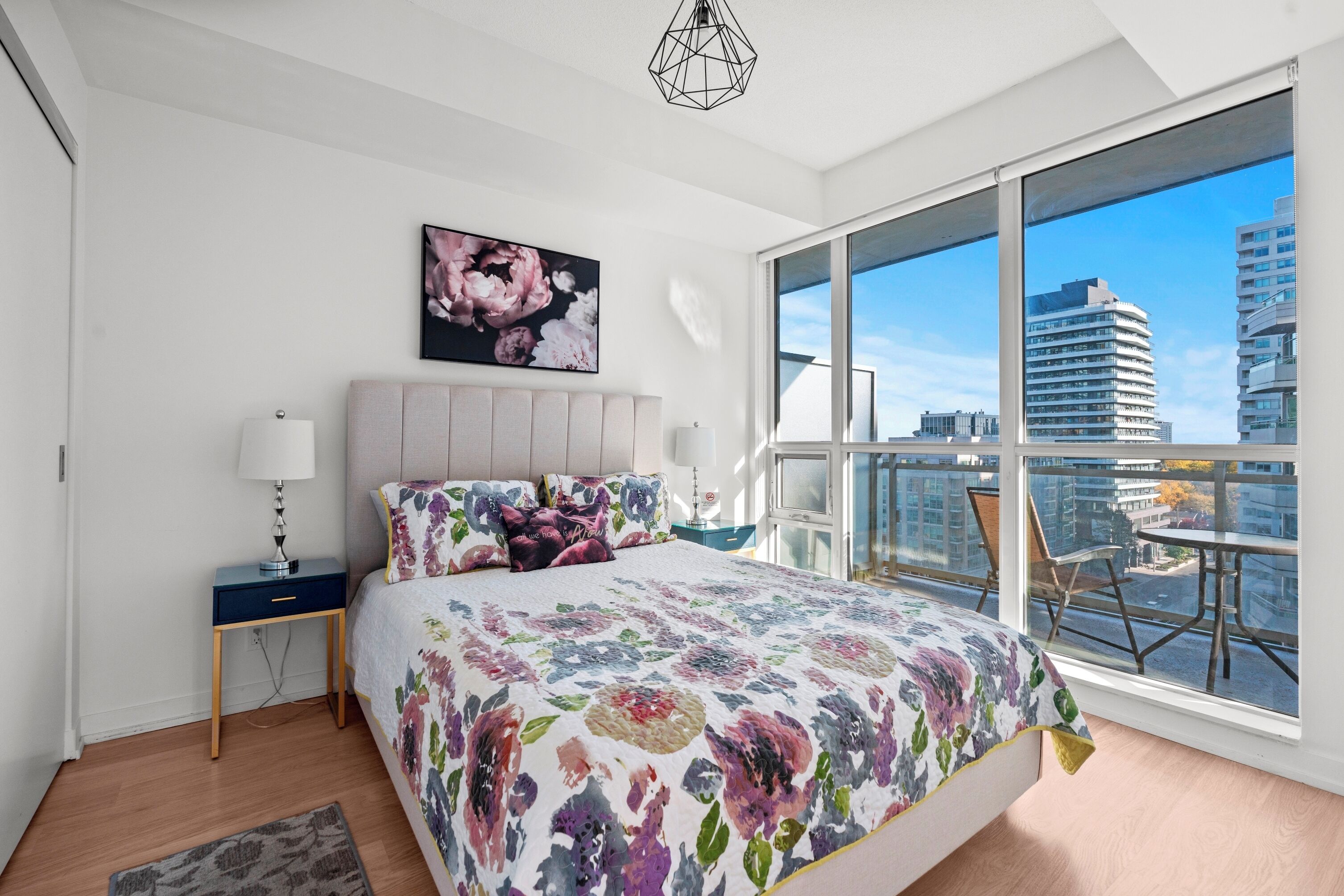 Un dormitorio moderno con una cama grande, una mesita de noche, una lámpara y vistas de la ciudad a través de amplias ventanas.