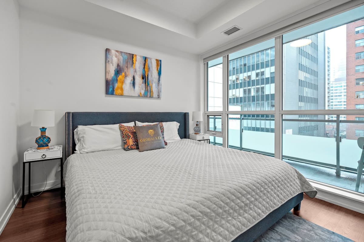 Ein modernes Hotelzimmer mit einem großen Bett, einem Nachttisch, einer Lampe, einem Gemälde und Blick auf die Stadt durch große Fenster.