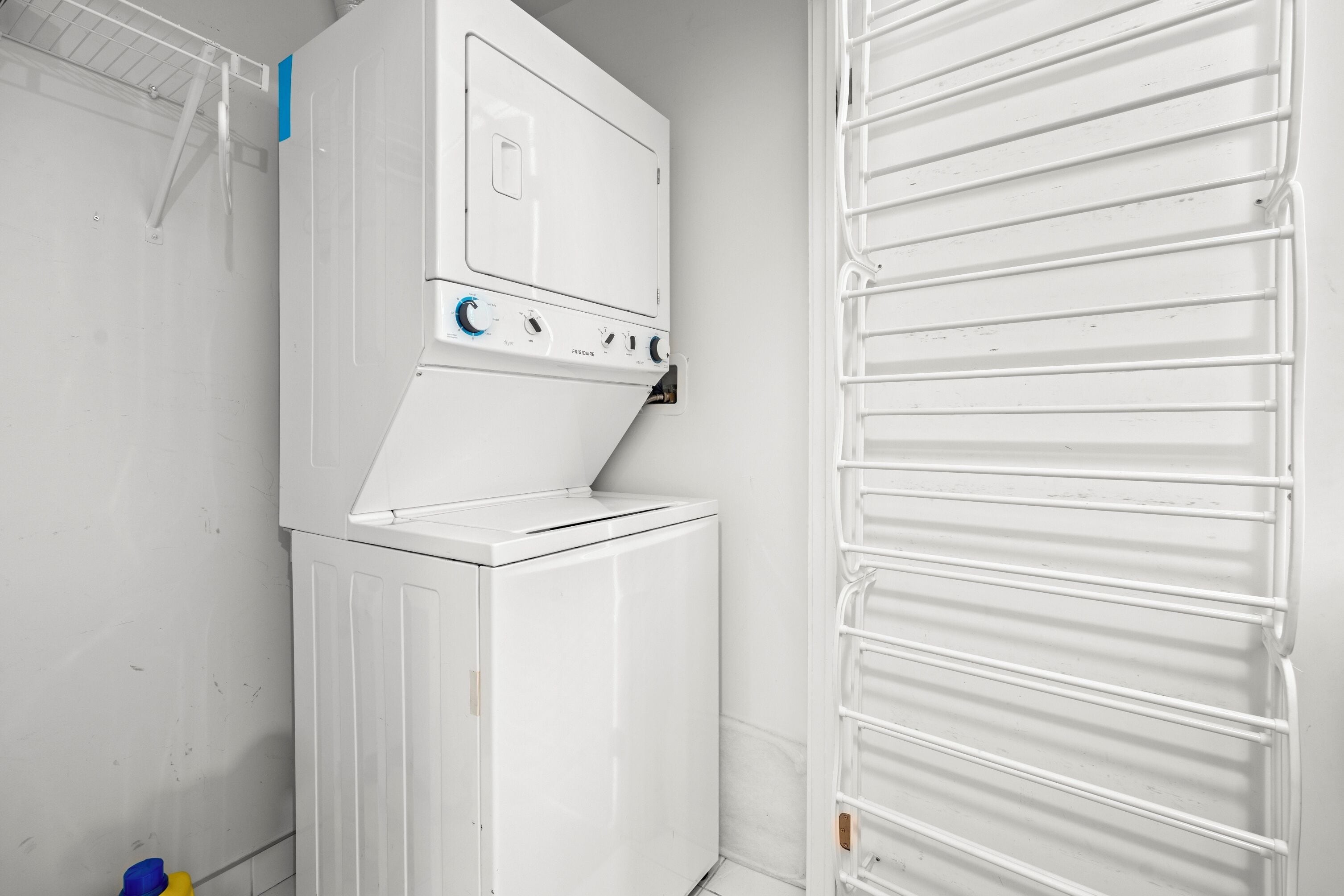 A compact laundry room with a stacked washer and dryer, a metal drying rack, and a small shelf with cleaning supplies.