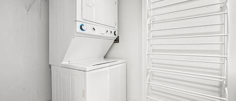 A compact laundry room with a stacked washer and dryer, a metal drying rack, and a small shelf with cleaning supplies.