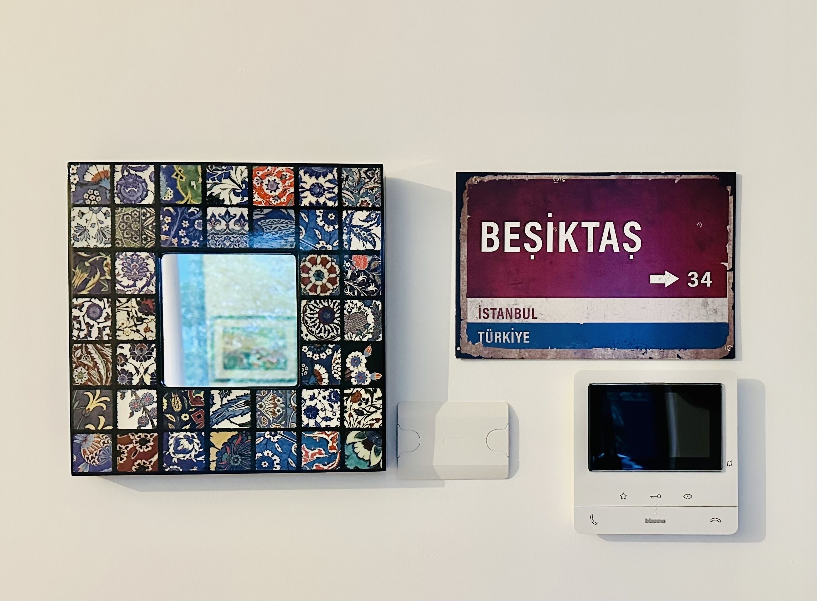 Eine Wand mit einem Mosaik-Wandbild, einem Schild mit der Aufschrift 'BESİKTAS' und einem Pfeil, der auf die Zahl 34 zeigt, sowie ein Thermostat.
