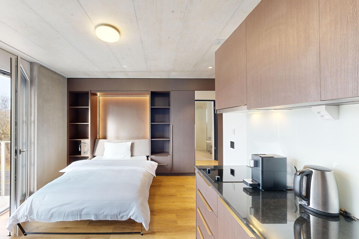 A modern bedroom with a large bed, built-in shelving, a kitchen area with a kettle, and a balcony with a railing.