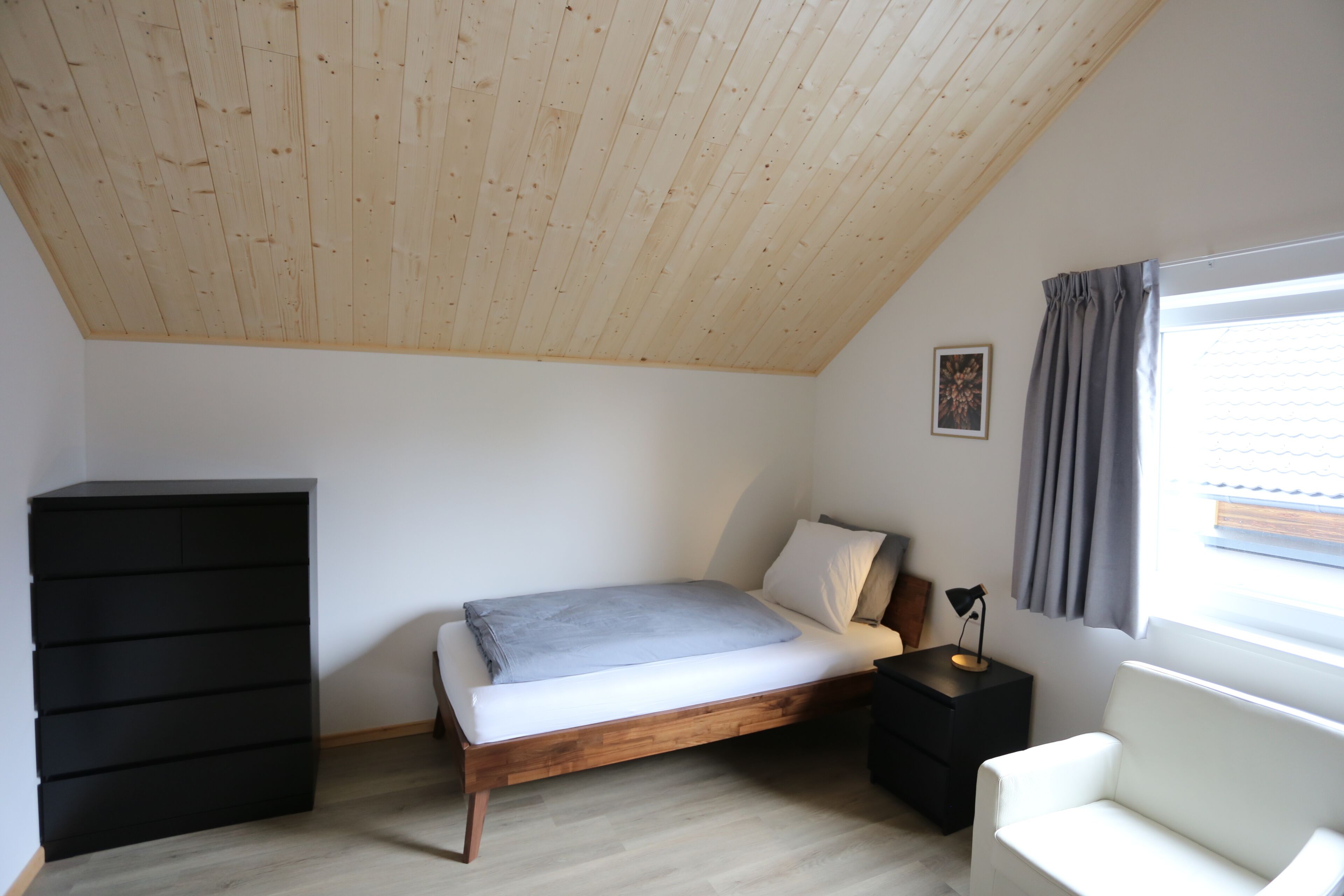 A bedroom with a slanted ceiling, a bed, a dresser, a nightstand, a lamp, a window with curtains, and a picture on the wall.