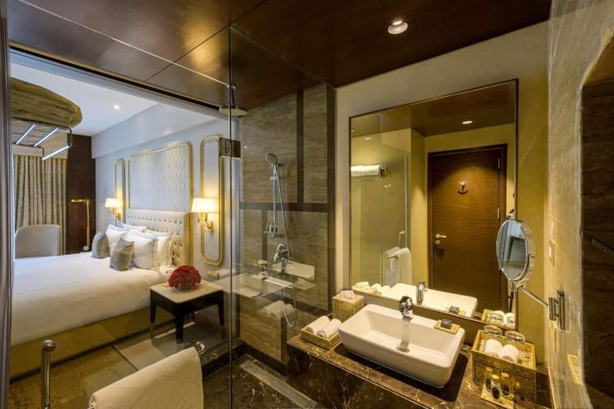 A modern hotel room with a glass-enclosed shower, a large bed, a vanity with a mirror, and a separate bathroom.